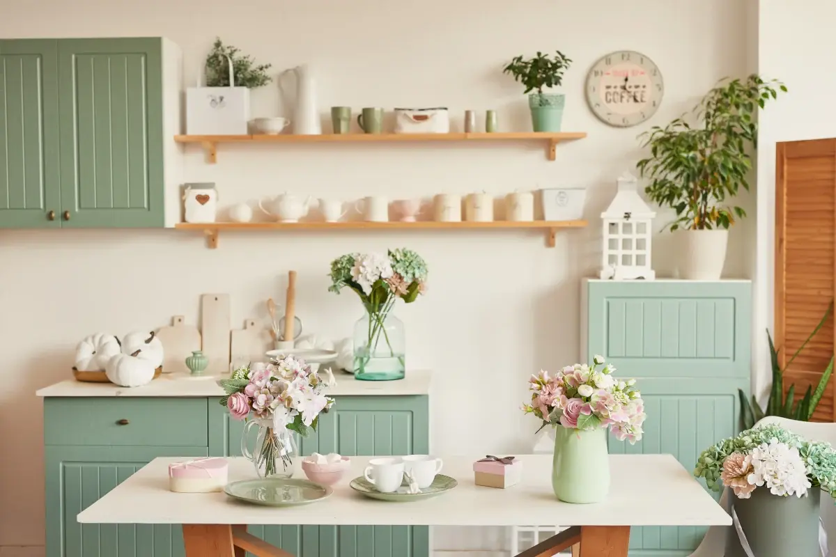 Udekorowany stół wazonem z kwiatami na tle mebli w kolorze zieleni oraz bieli z elementami drewna.