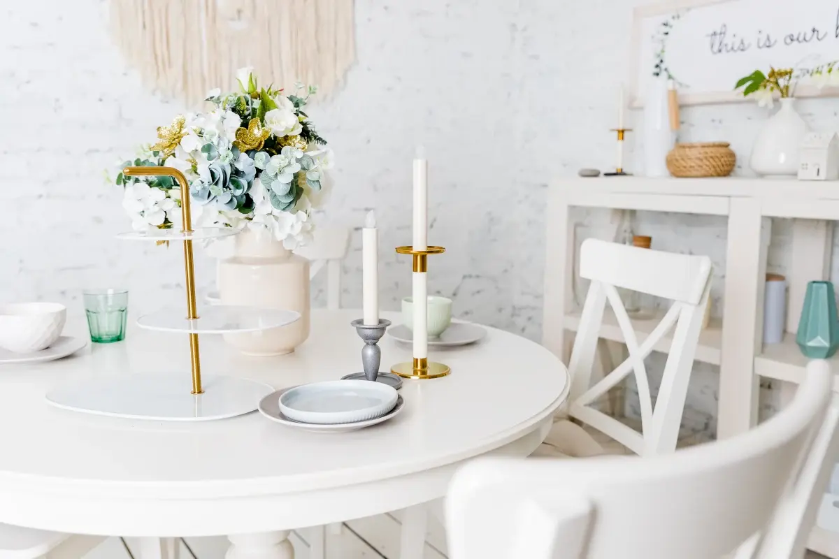 Biały stół z krzesłami z zastawą oraz udekorowany wazonem z kwiatami oraz świecznikami.