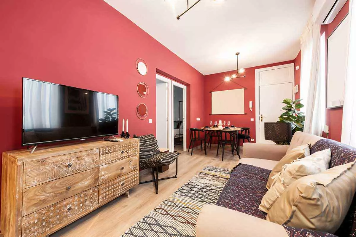 Salon z pomalowanymi na czerwono ścianami. Komoda drewniana, na której stoi telewizor.. Białe drzwi oraz listwy w pokoju.