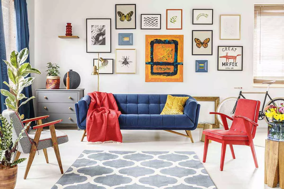 Salon z niebieską kanapą. Czerwony fotel oraz koc. Na ścianie dużo obrazów.