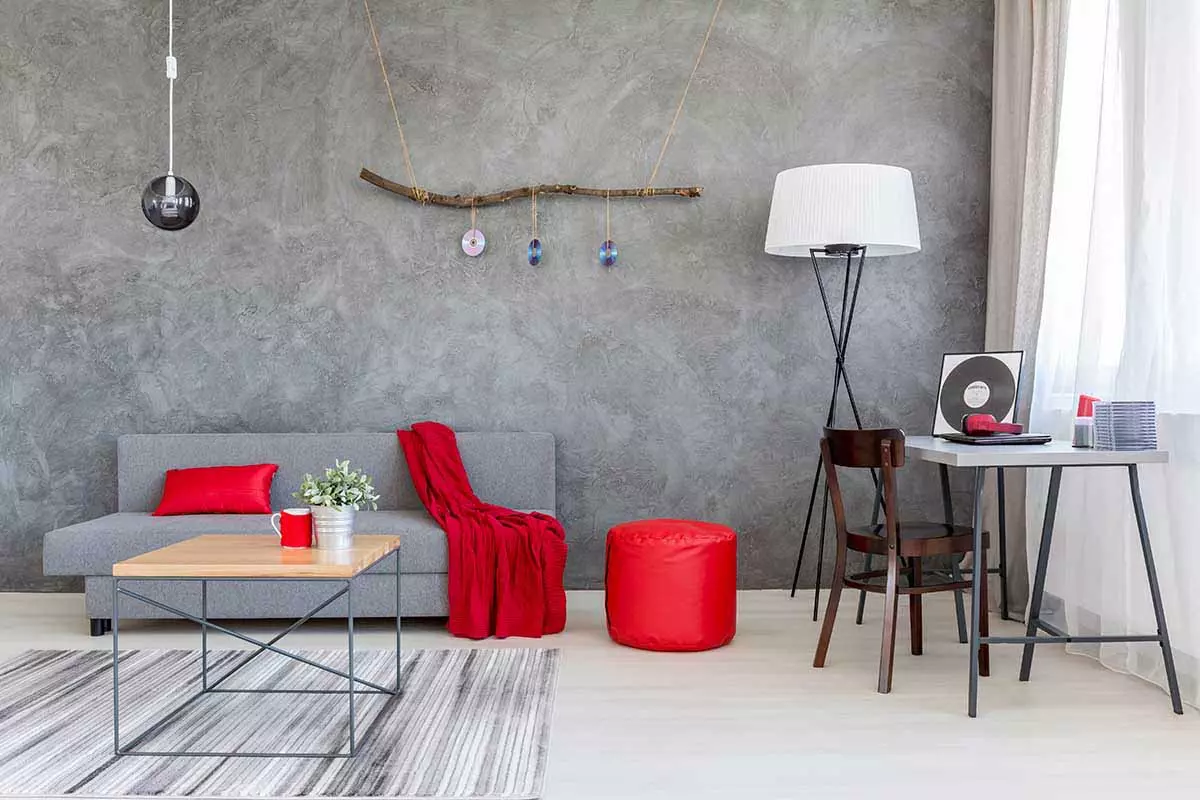 Szara kanapa na tle betonu na ścianie. Poduszka, koc oraz pufa w kolorze czerwonym.