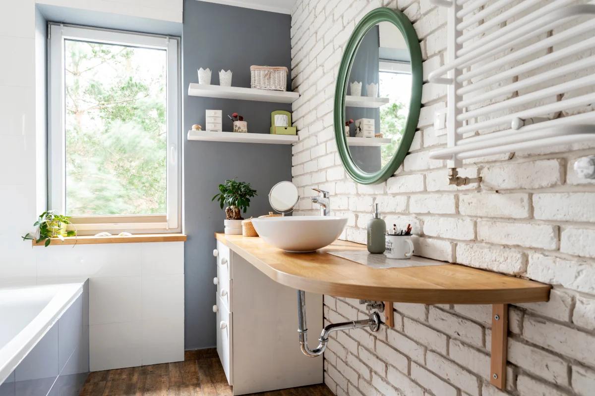 Łazienka w kolorze białym z ceglaną ścianą, drewnianym blatem, umywalką i wiszącym, okrągłym lustrem.