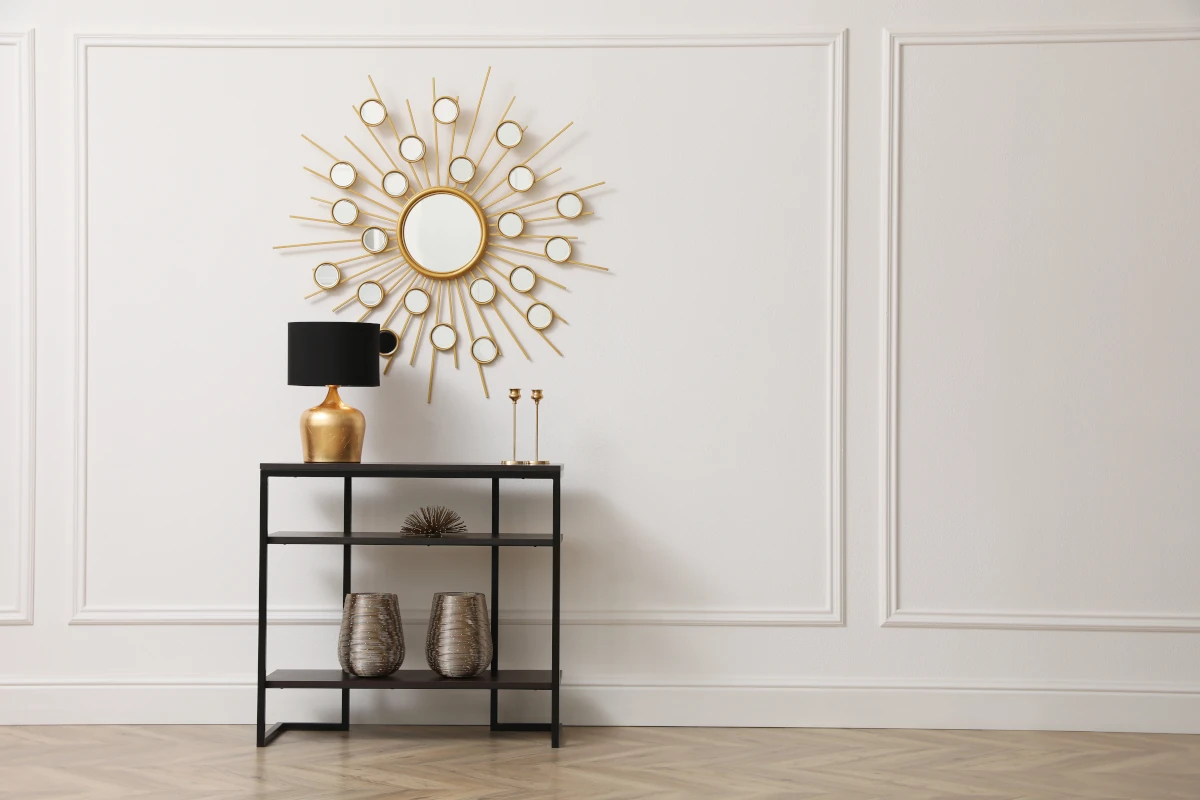 Czarna konsola stojąca pod białą ścianą. Nad konsolą lustro w kształcie słońca. Na konsoli lampa oraz wazony.