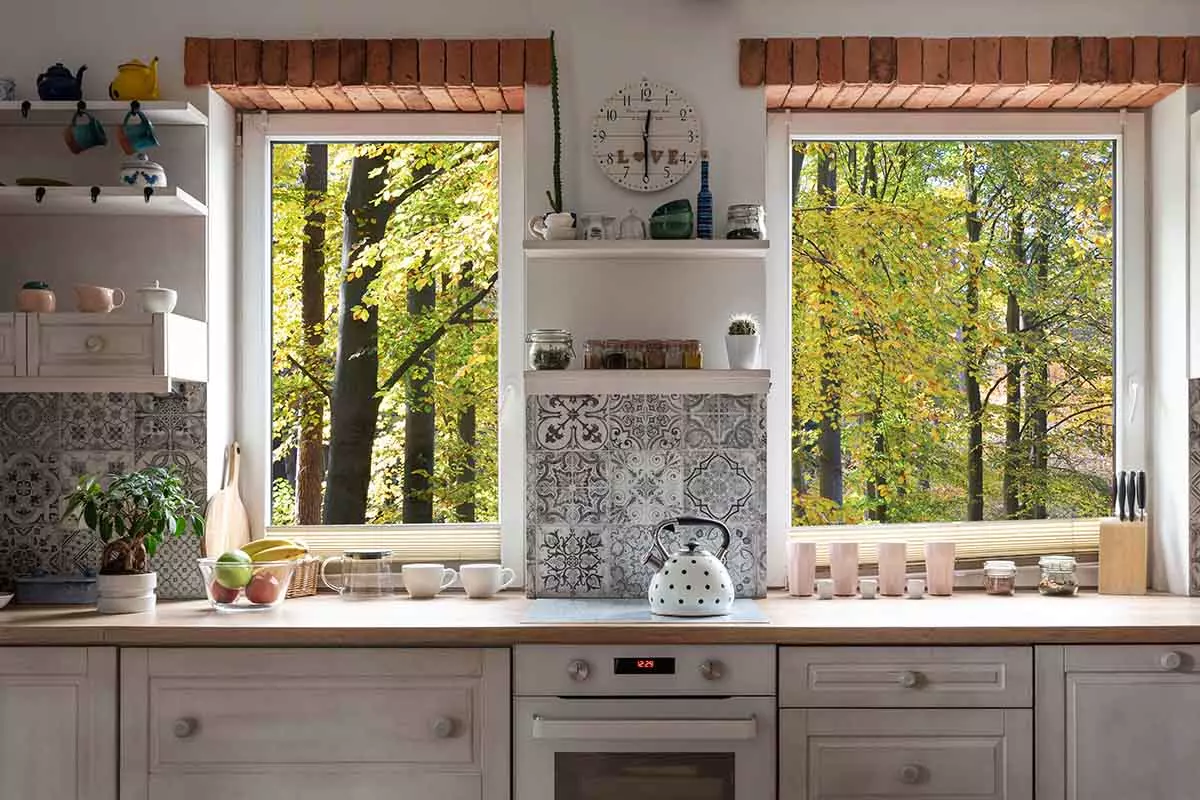 Meble kuchenne pod oknem. Fronty beżowe, blat drewniane. Nad blatem są płytkim ze wzorem oraz półki z dekoracjami.
