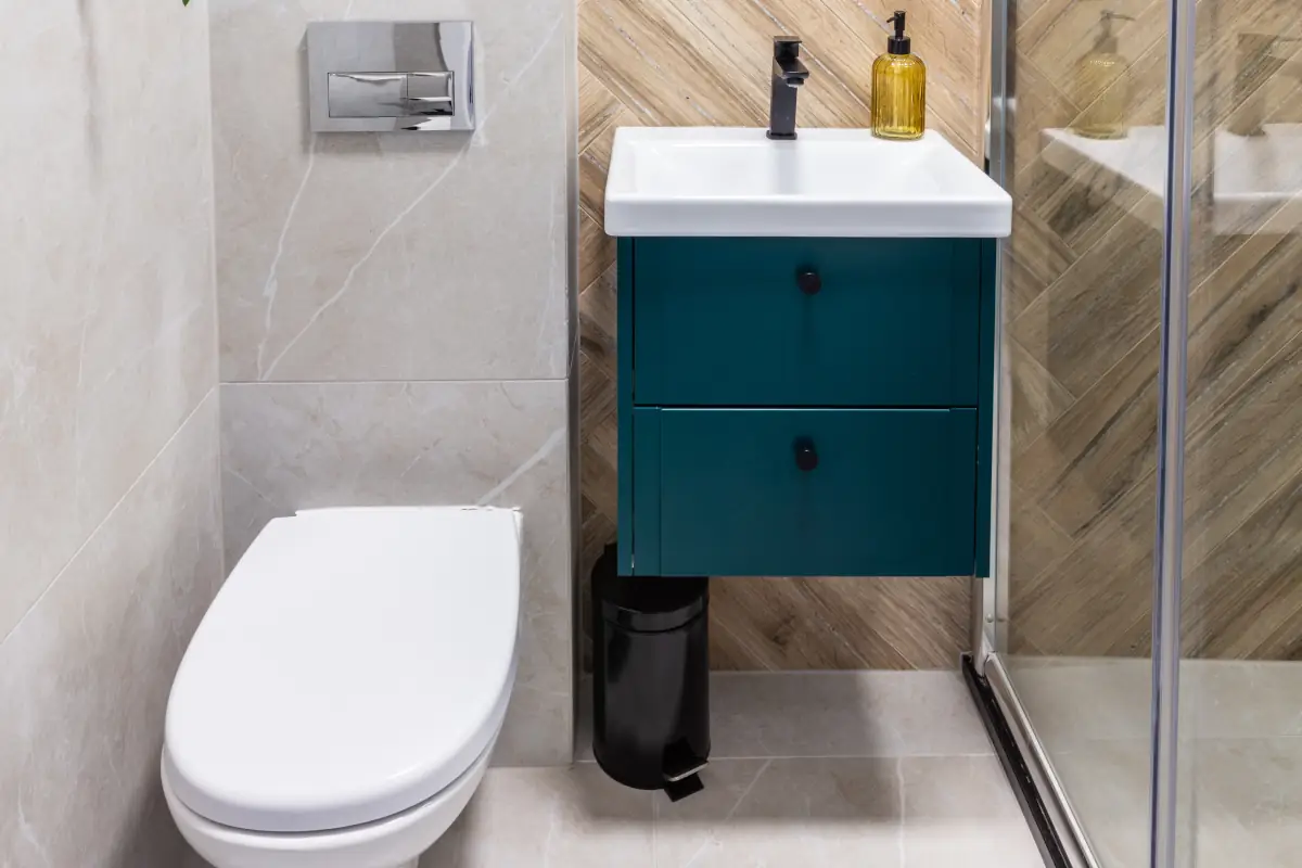 Mała łazienka w połączeniu kolorów: bieli, szarości oraz drewna. Lustro wiszące nad umywalka na niebieskiej szafce obok miski wc.
