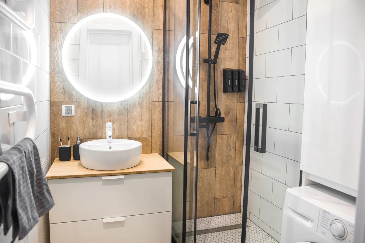 Mała łazienka z płytkami w kolorze bieli oraz imitujące drewno. Kabina prysznicowa obok umywalki. Czarna armatura oraz akcesoria.