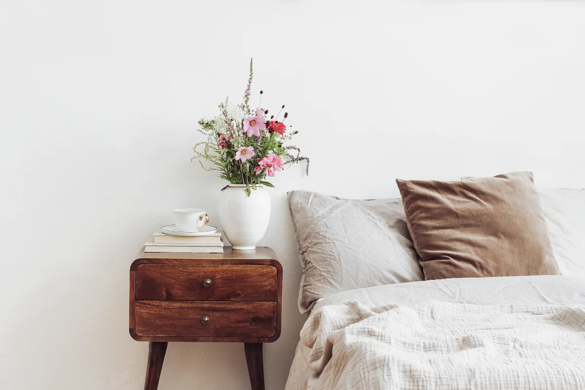 Poduszki oraz narzuta w kolorach ziemi. Obok łóżka stoi starodawny, drewniany stolik nocny. Na nim leżą książki oraz stoi wazon z kwiatami.