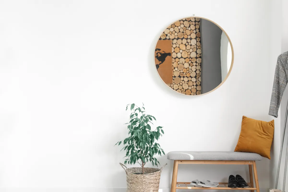 Drewniana ławeczka pod ścianą obok wiklinowej osłonki z kwiatem. Powyżej ławeczki lustro.