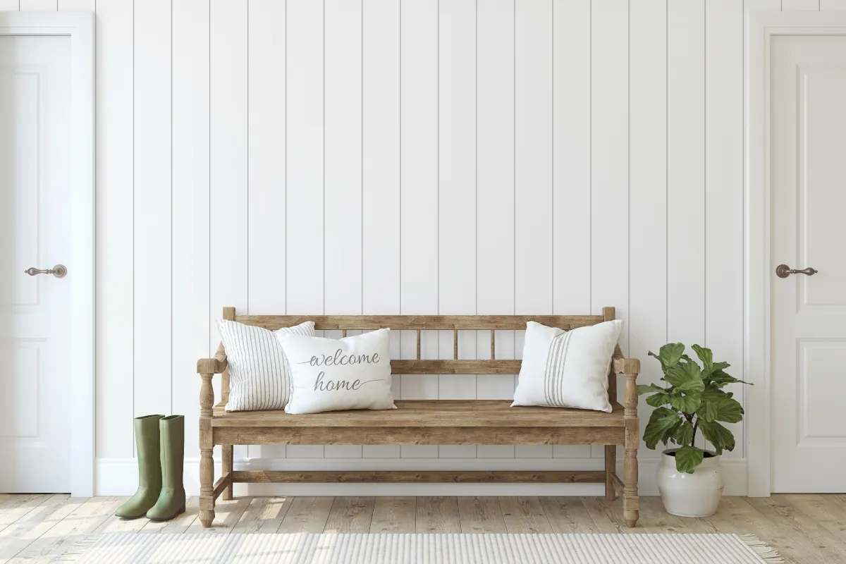 Drewniana ławka z poduszkami na tle białej ściany z paneli. Obok ławki donica z kwiatem oraz kalosze.
