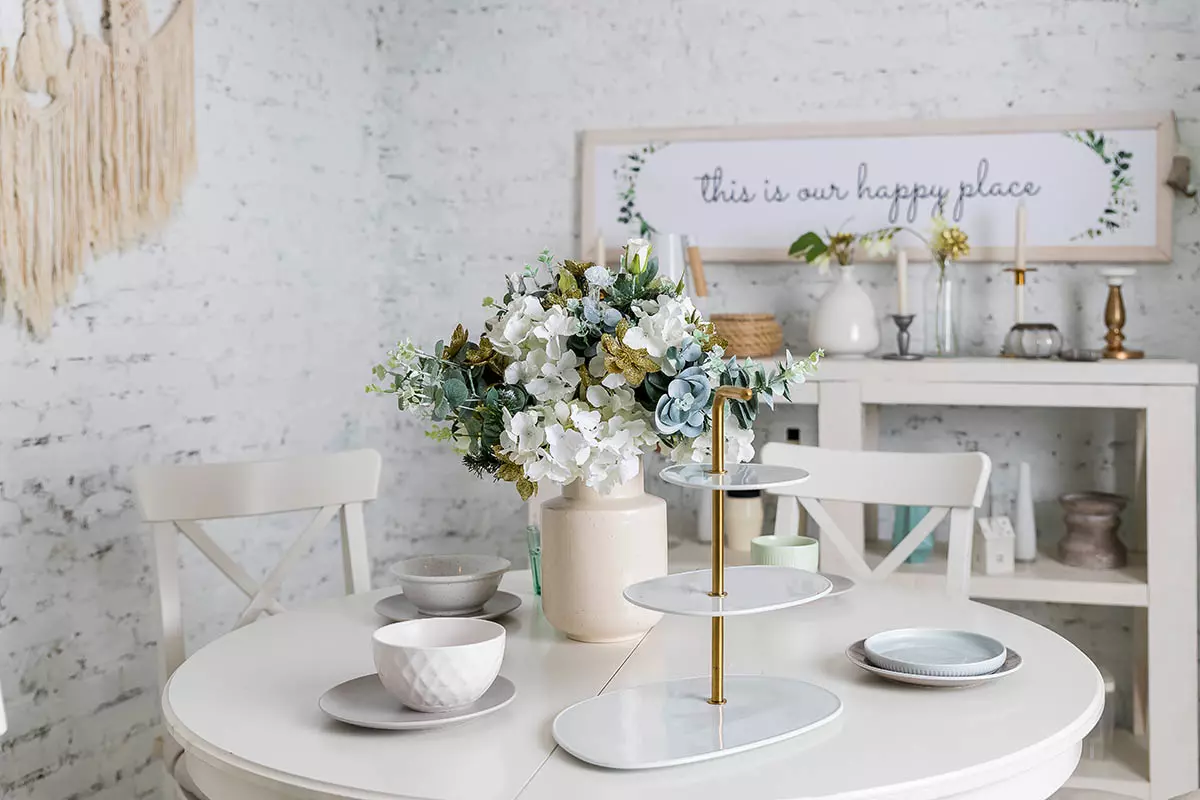 Pomieszczenie z naturalną cegłą na ścianie. Na stole stoi zastawa oraz wazon z kwiatami. W tle na regale różne akcesoria i dekoracje.