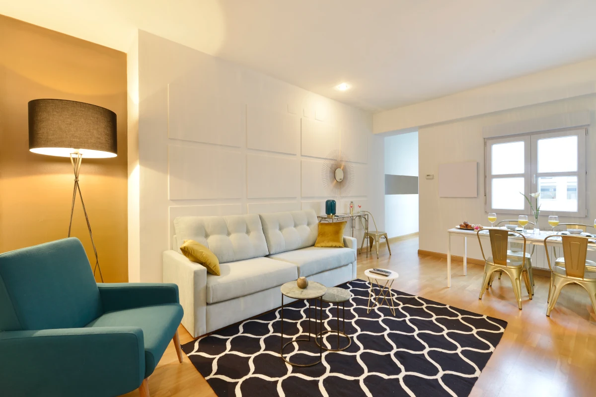 Pomieszczenie będące połączenie salonu oraz jadalni. Kanapa jasna na tle białej ściany. Zielony fotel na tle złotej ściany. Na podłodze dywan we wzory.