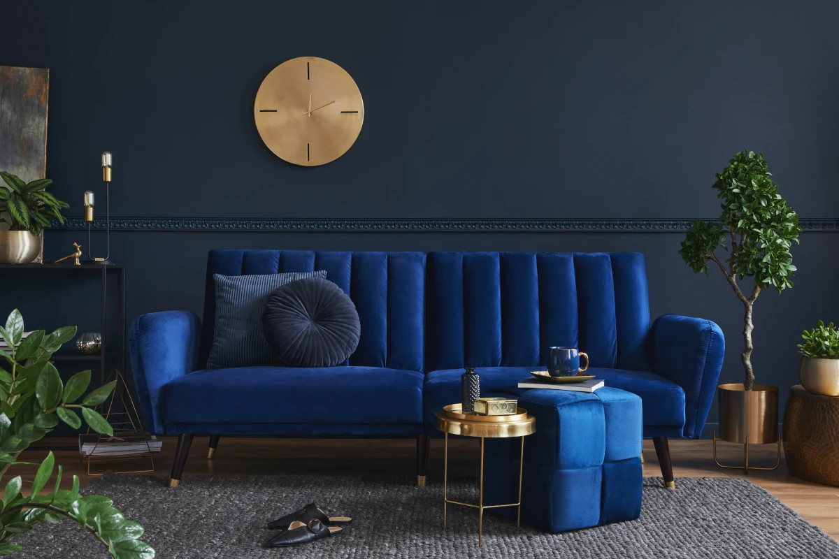 Niebieska kanapa na tle granatowej ściany. Wszelkie dodatki są w kolorze złota, co stanowi kontrast.