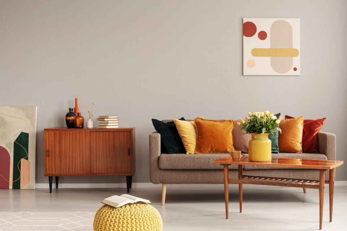 Sofa z kolorowymi poduszkami stojąca przy stoliku z wazon. Obok sofy stoi szafka. Na ścianie obrazy.