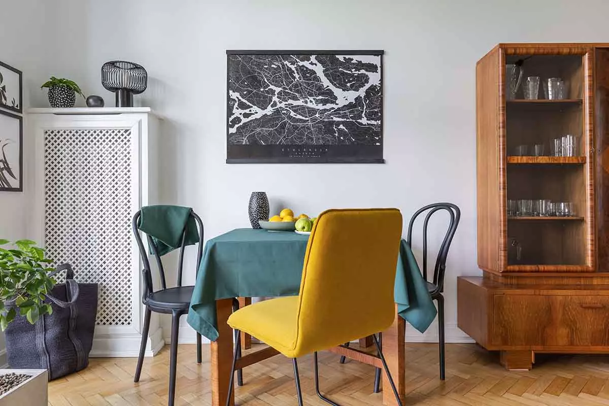 Stół w pokoju z zielonym obrusem obok żółtego krzesła. Obok witryna ze szklanymi dekoracjami.