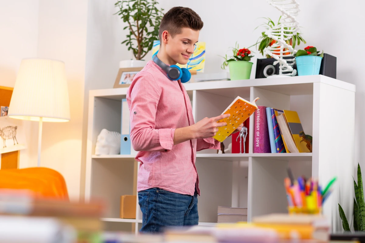 Chłopak w różowej koszuli stojący na środku pokoju i trzymający książkę w dłoniach. Na szyi ma zawieszone słuchawki. W tle znajduje się regał na książki i inne dekoracje.