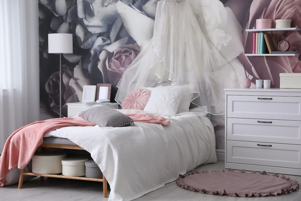 Łóżko z drewnianą konstrukcją z białą kołdrą oraz poduszkami. Narzuta na łóżku różowa. Na ścianie za łóżkiem róże.