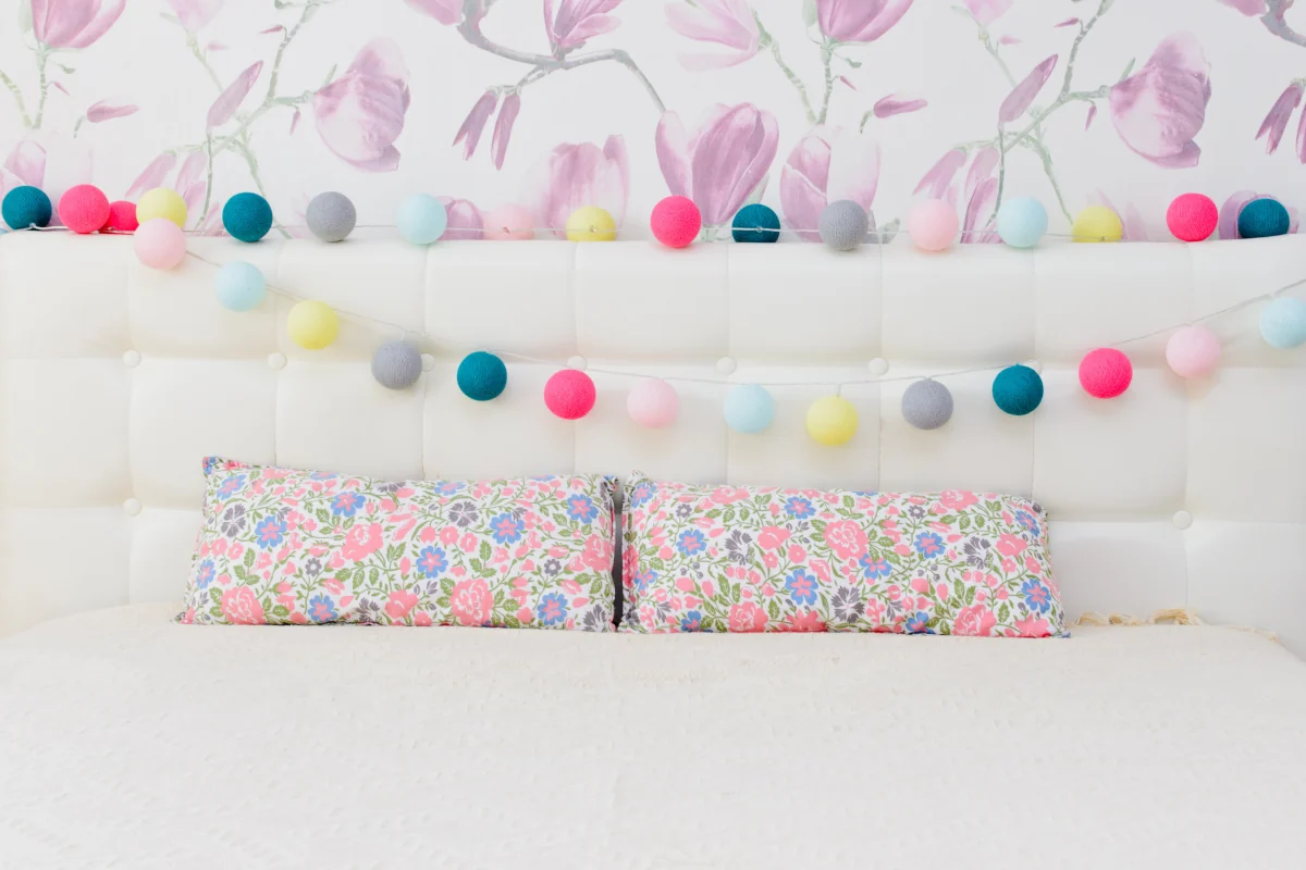Łańcuch świetlny zawieszony na oparciu łóżka. Kolorowe poduszki w kwiaty.