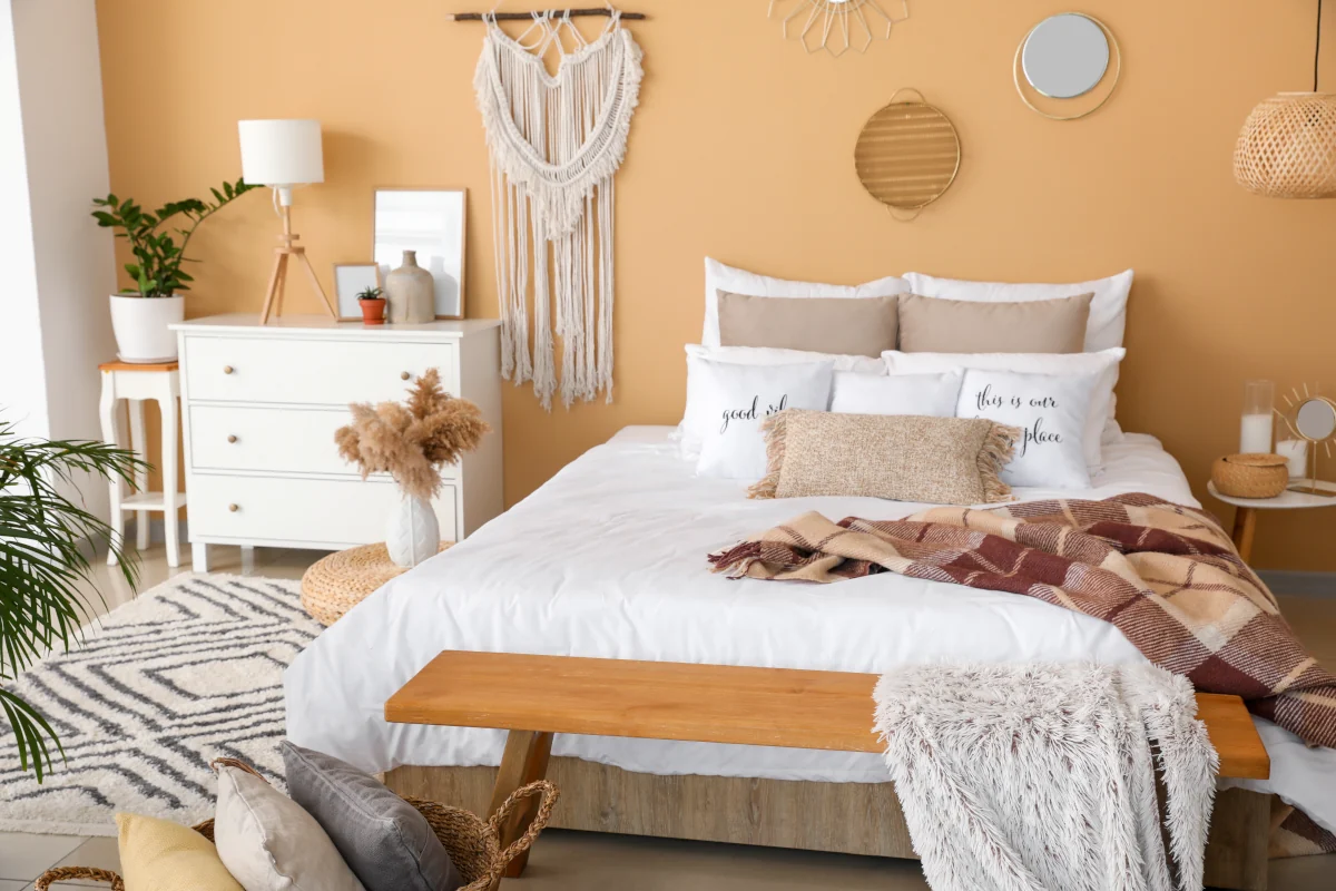 Łóżko z białą kołdrą oraz poduszkami w kolorze ziemi. ściana za łóżkiem pomarańczowa z makramą. Obok łóżka stoi komoda z dekoracjami.
