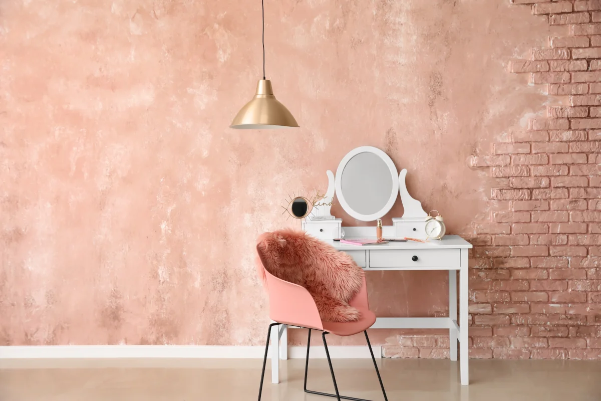 Toaletka biała na tle ściany z różowym tynkiem oraz cegłą. Fotel przy toaletce różowy.