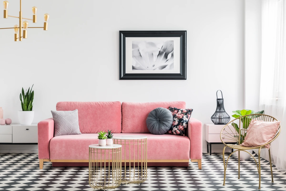 Różowa kanapa na tle białej ściany. Stolik kawowy, krzesła oraz inne dekoracje w stylu glamour.