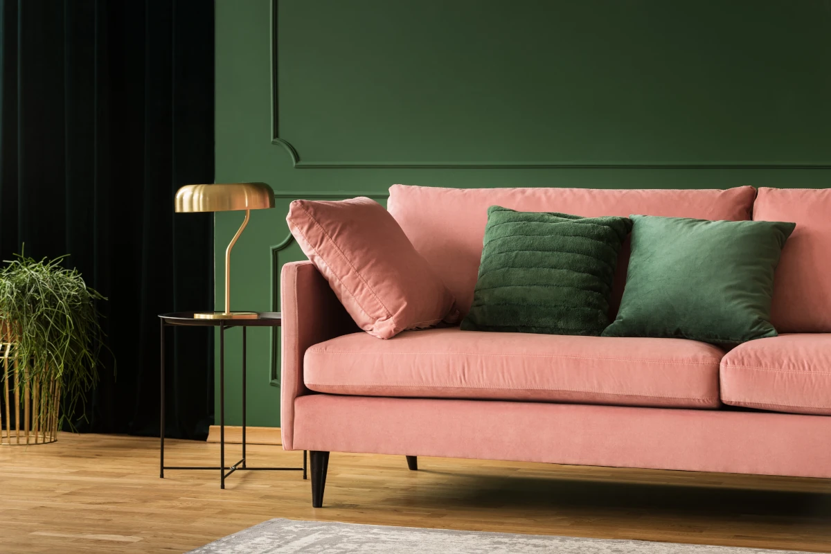 Sofa w różowych odcieniach z zielonymi poduszkami na tle zielonej ściany. Dodatki w postaci firan w kolorze czarnym, a na stoliku lśniąca lampa.