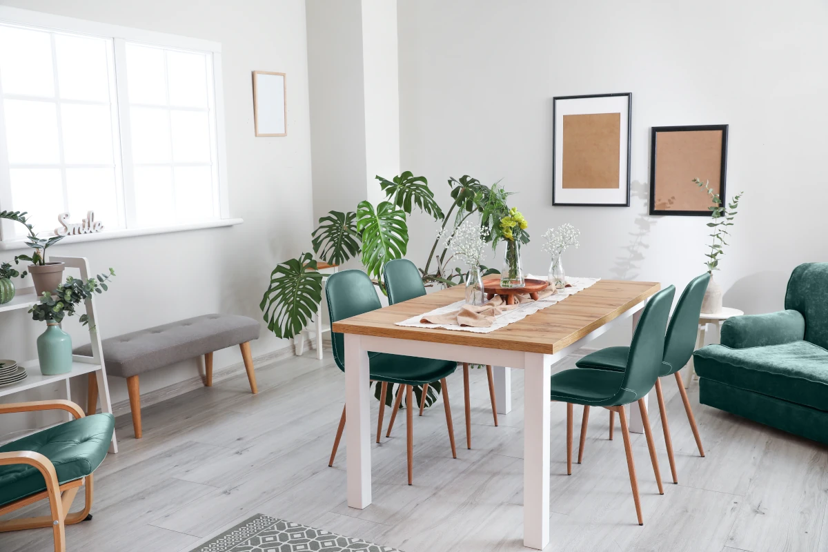 Jadalnia z białym stołem i blatem drewnianym oraz z krzesłami z zielonymi siedziskami. Duże okno daje sporo światła dla roślin znajdujących się w pomieszczeniu.