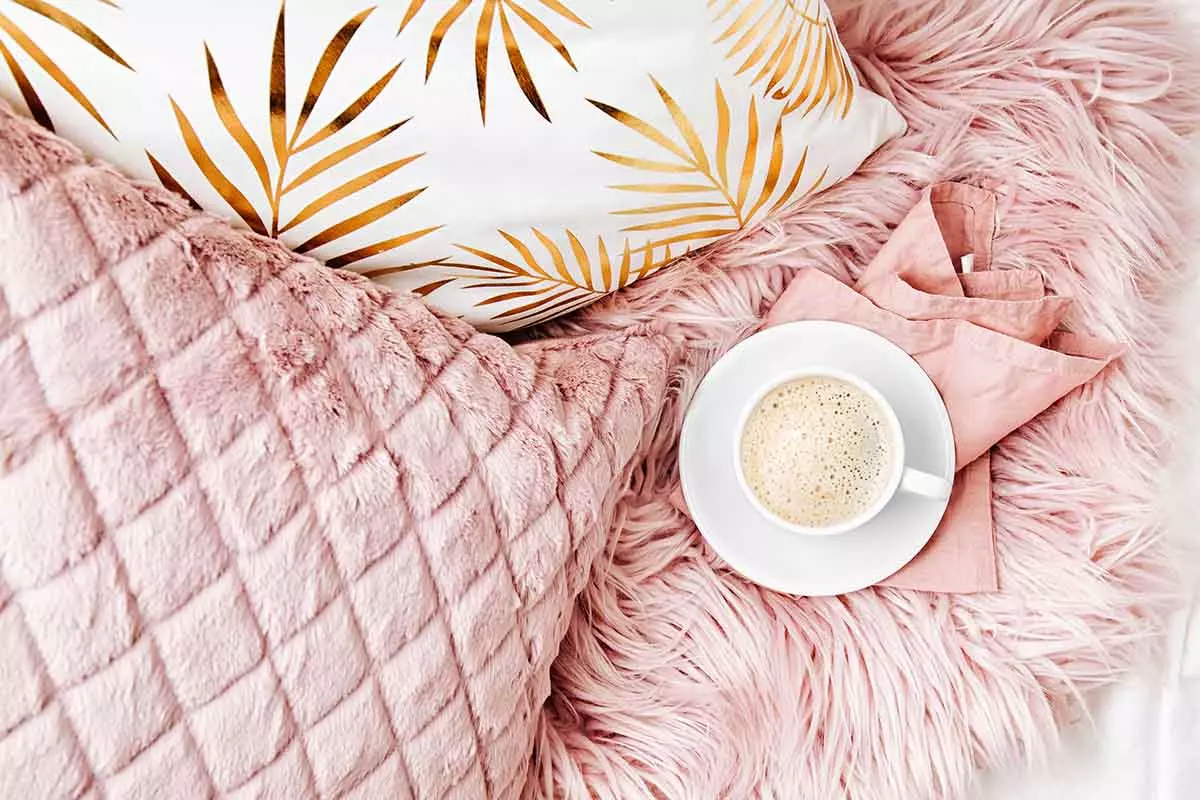 Postawiona kawa na poduszkach. Jedna poduszka i narzuta w kolorze różu. Druga poduszka ze złotymi akcentami liści.