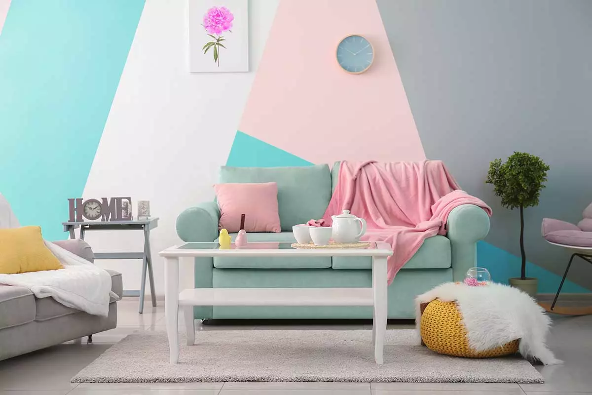 Sofa w kolorze szałwiowym z poduszkami oraz narzutami w kolorze różu. Obok druga szara kanapa. Ściana pomalowana na różne kolory w geometryczne kształty.