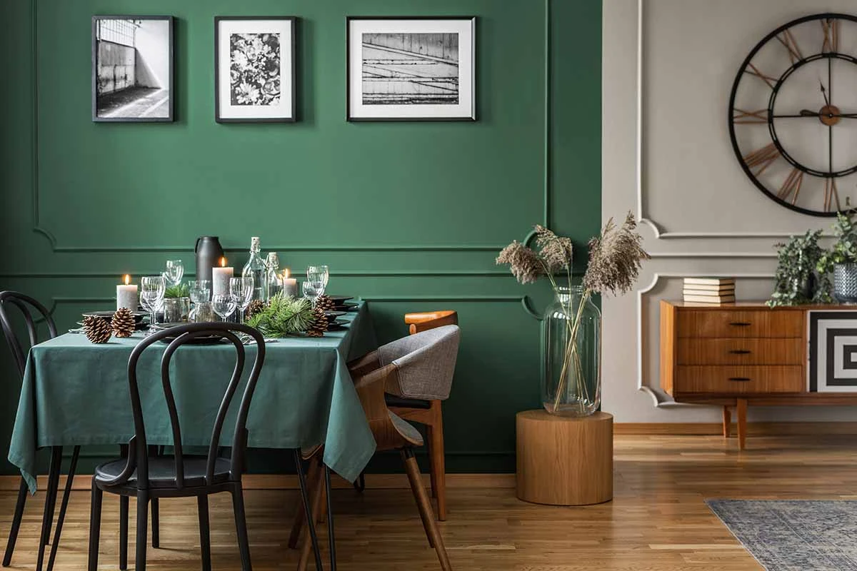 Zastawiony stół z zielonym obrusem oraz krzesłami. Wnętrze z ścianami w kolorze zielonym oraz szarym. Podłoga w kolorze drewna.