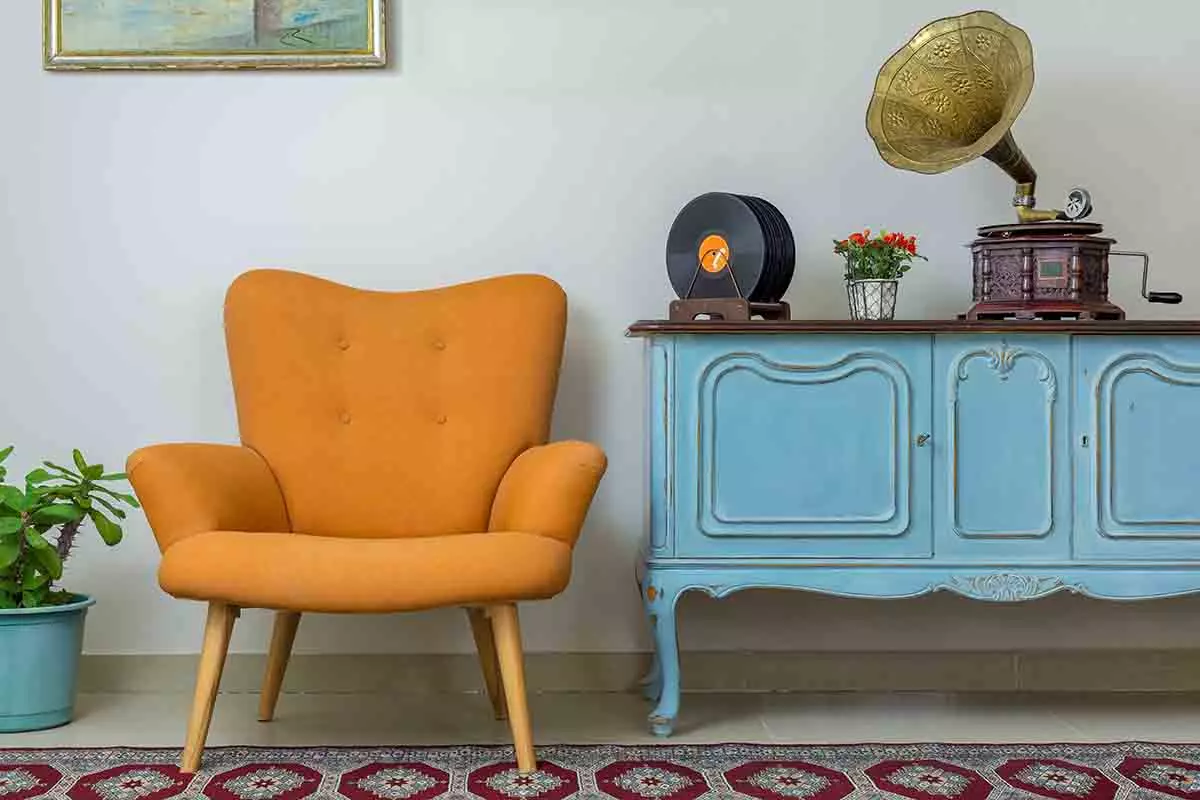 Pomarańczowy fotel obok niebieskiej szafki. Na szafce postawiono gramofon oraz płyty winylowe.