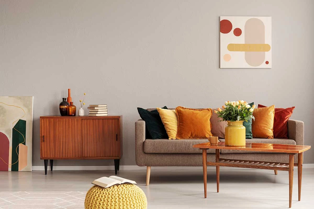 Sofa z kolorowymi poduszkami. Stolik z żółtym wazonem i kwiatami. Nad sofą zawieszony obraz.