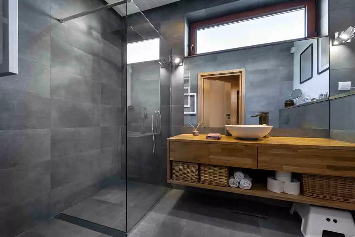 Łazienka w prysznicem typu walk-in. Drewniana szafka pod umywalkę. Płytki w kolorze szarym.