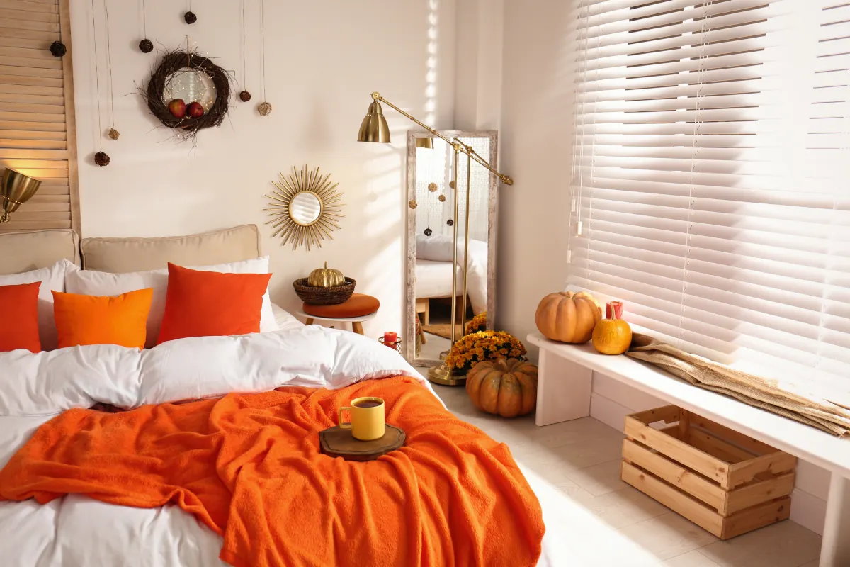 Łóżko w sypialni z poduszkami oraz narzutami w kolorze pomarańczowym. Dynie stojące pod oknem.