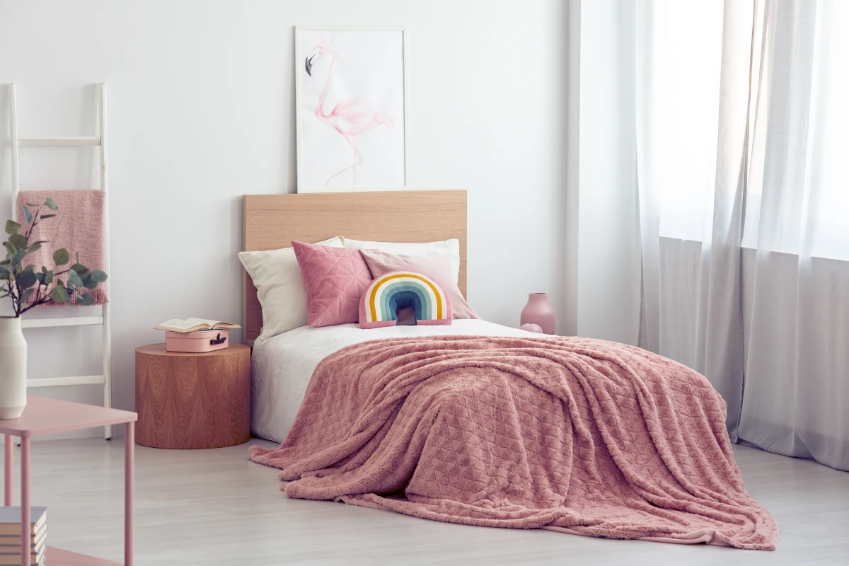 Pokój dziewczynki z łóżkiem z poduszkami oraz narzutą w kolorze różowym. Poduszka w kształcie tęczy.