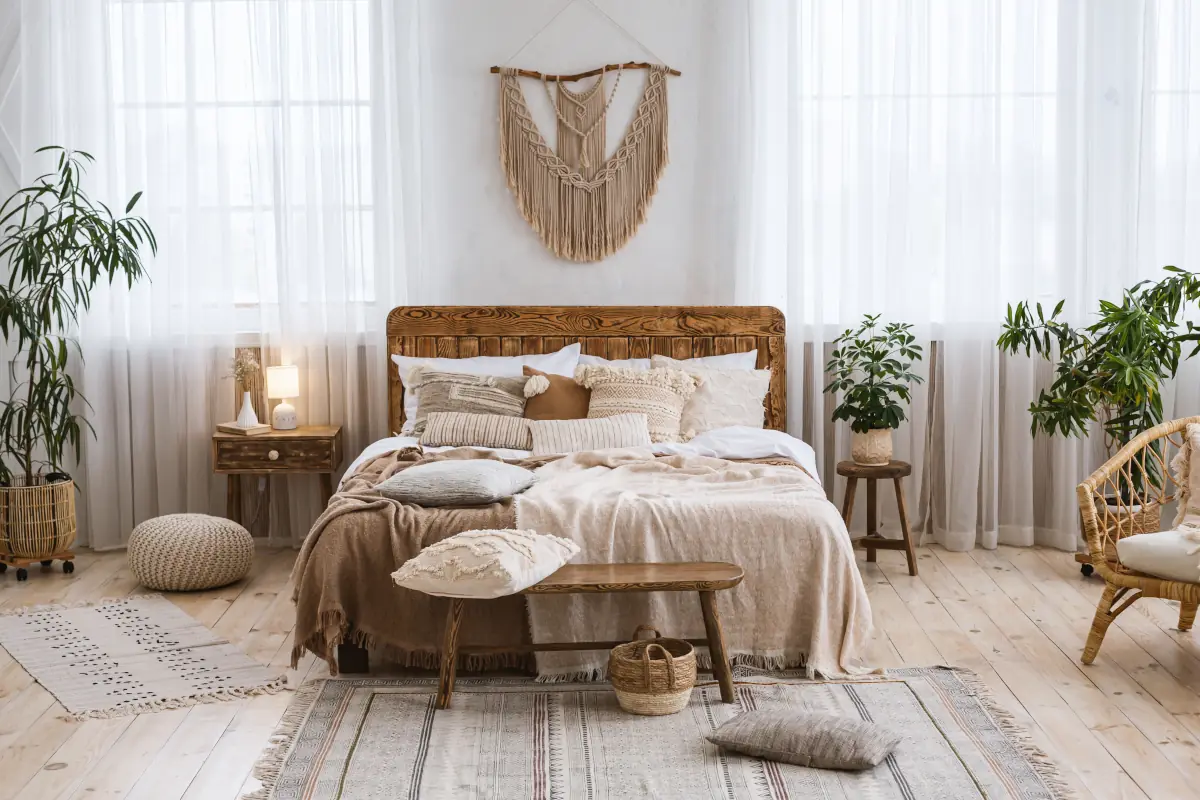 Łóżko w sypialni z poduszkami oraz narzutami w kolorze beżowym oraz brązowym. Nad łóżkiem znajduje się łapacz snów. Kwiaty również są dekoracje tego pomieszczenia.